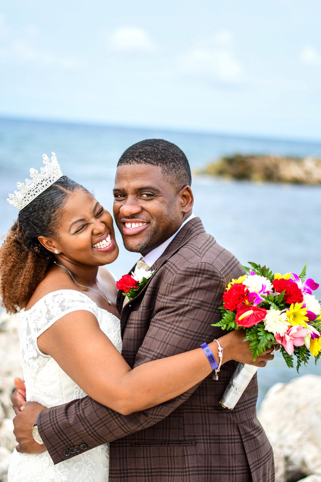 A bride in a tiara and her groom embrace and laugh by the sea