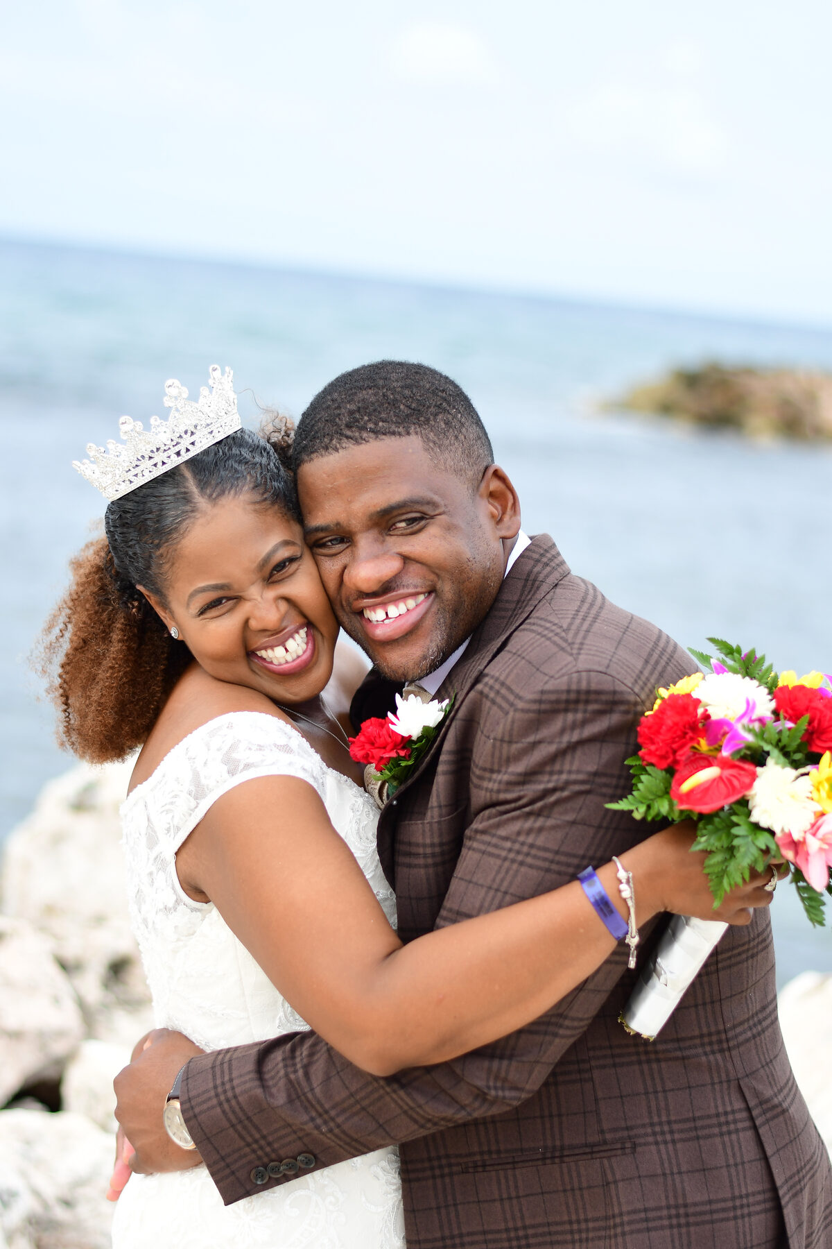 A bride and groom smile together holding a bright bouquet by the sea