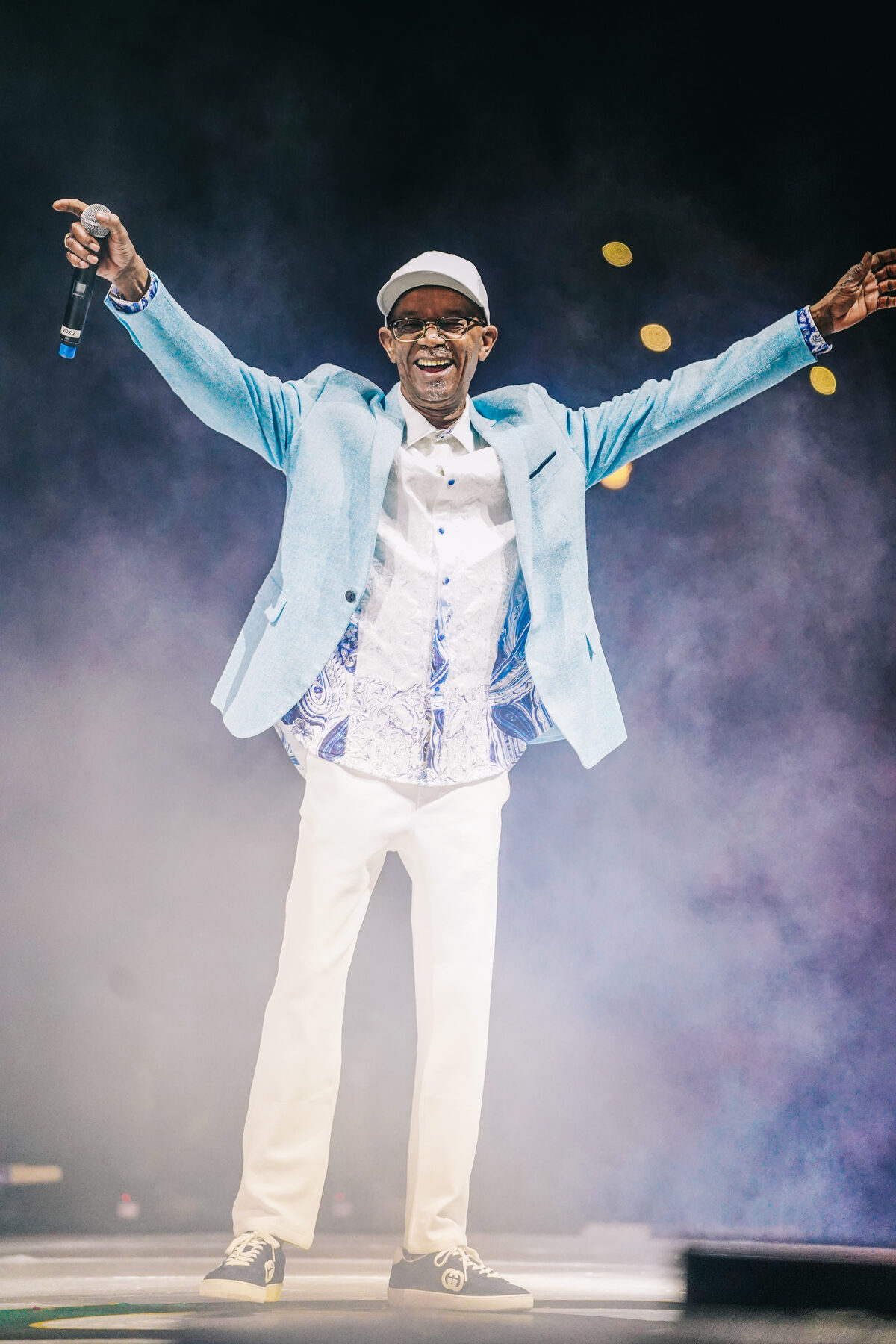 An artist in a white suit performs arms-wide in stage smoke and spotlight