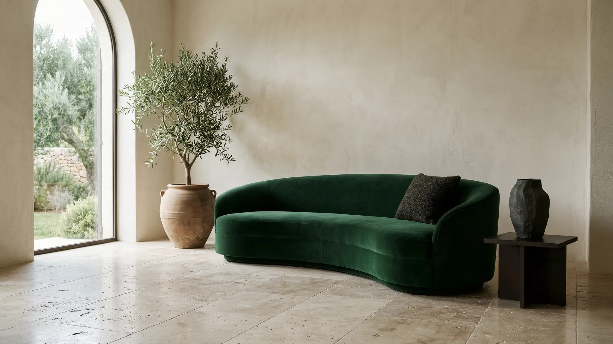 A curved emerald velvet sofa and a potted olive tree in a pale plaster room.