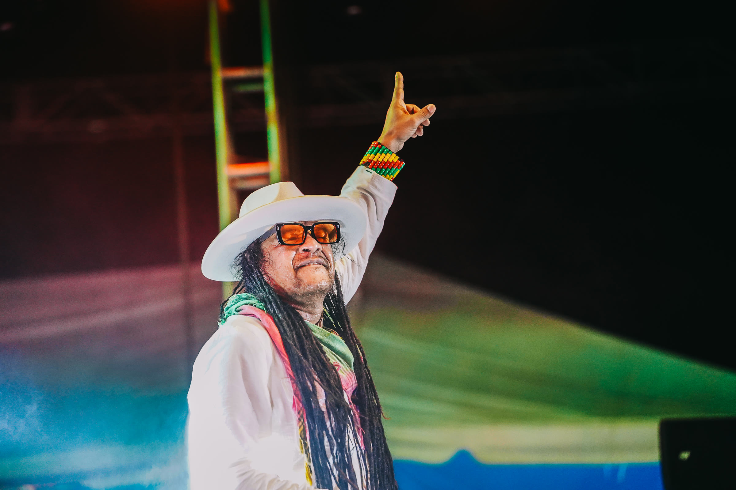 A reggae artist in a white hat and orange shades raises his hand on the Rebel Salute stage