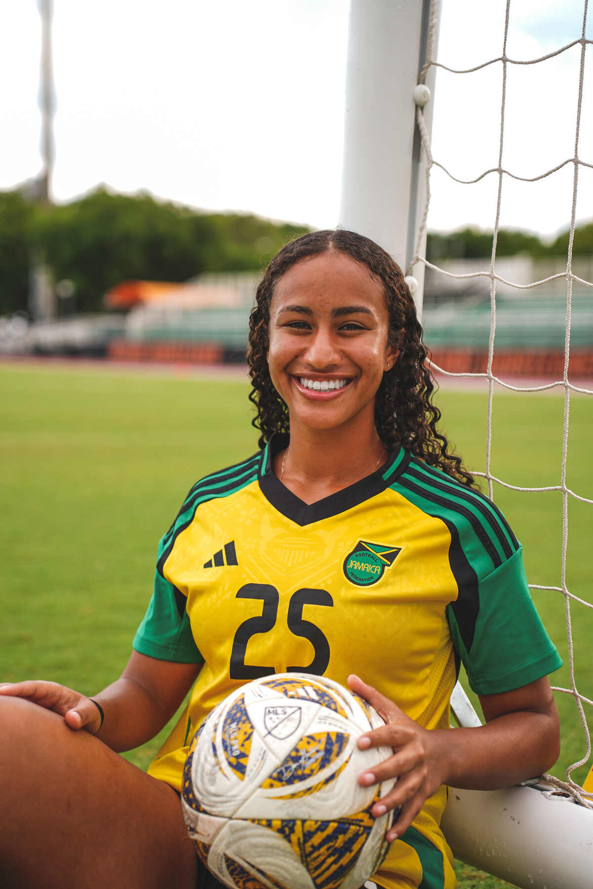 A young athlete in a Jamaica jersey holds a ball, smiling in the sun